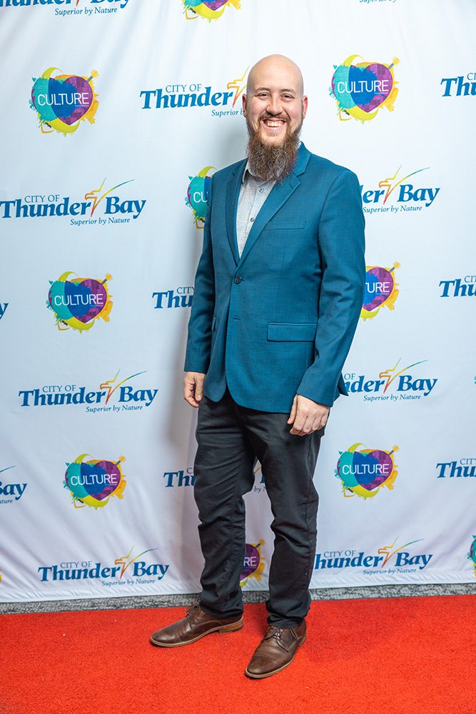 Bearded man in teal blue blazer standing confidently on red carpet with City of Thunder Bay backdrop