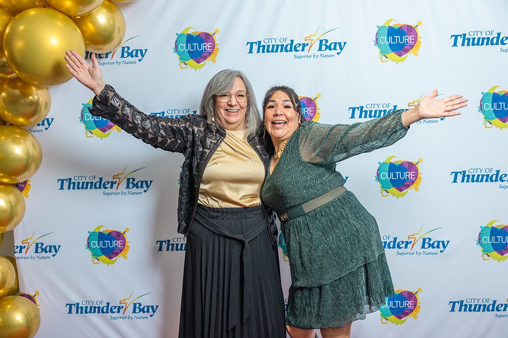 Two women with arms raised joyfully on red carpet with gold balloons at culture awards