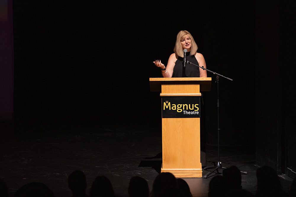 Woman in black dress speaking at wooden podium on dark stage at Magnus Theatre
