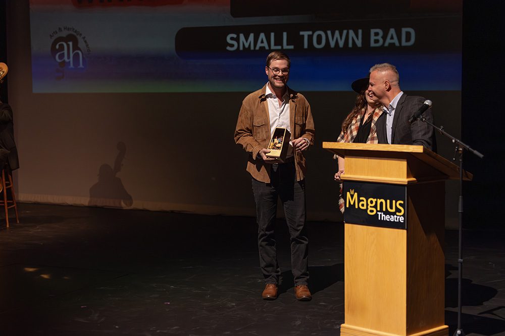 Award winner holding trophy on stage with presenters at Arts & Heritage Awards ceremony
