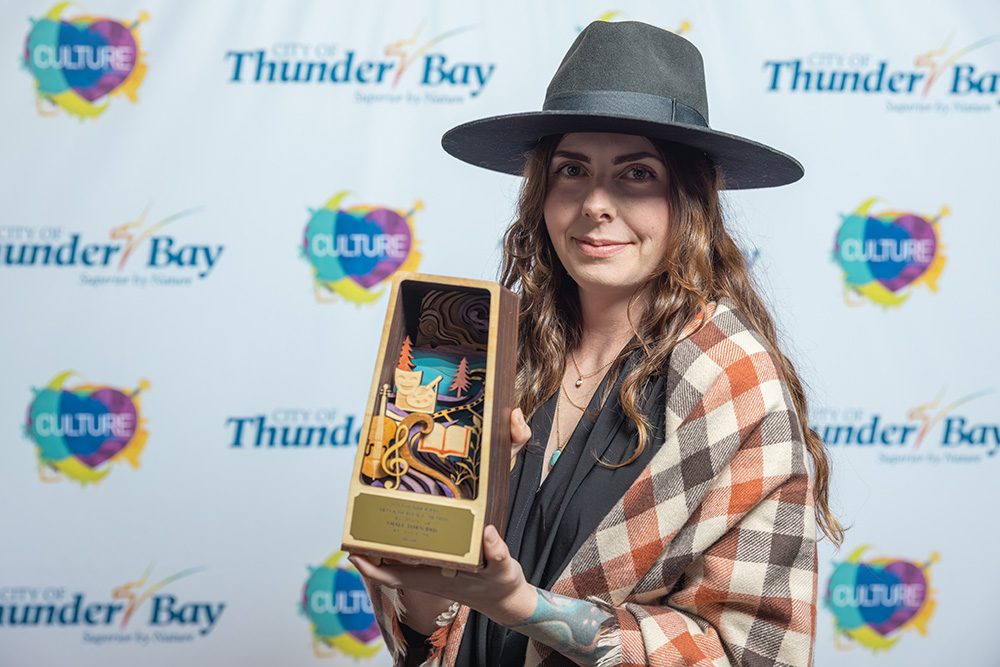 Woman in wide-brim hat and plaid blazer holding artistic award trophy on red carpet