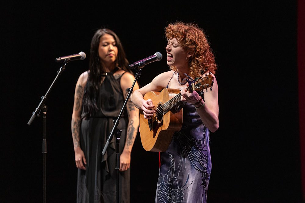 Two female musicians performing on stage, one with guitar one singing, in elegant dresses