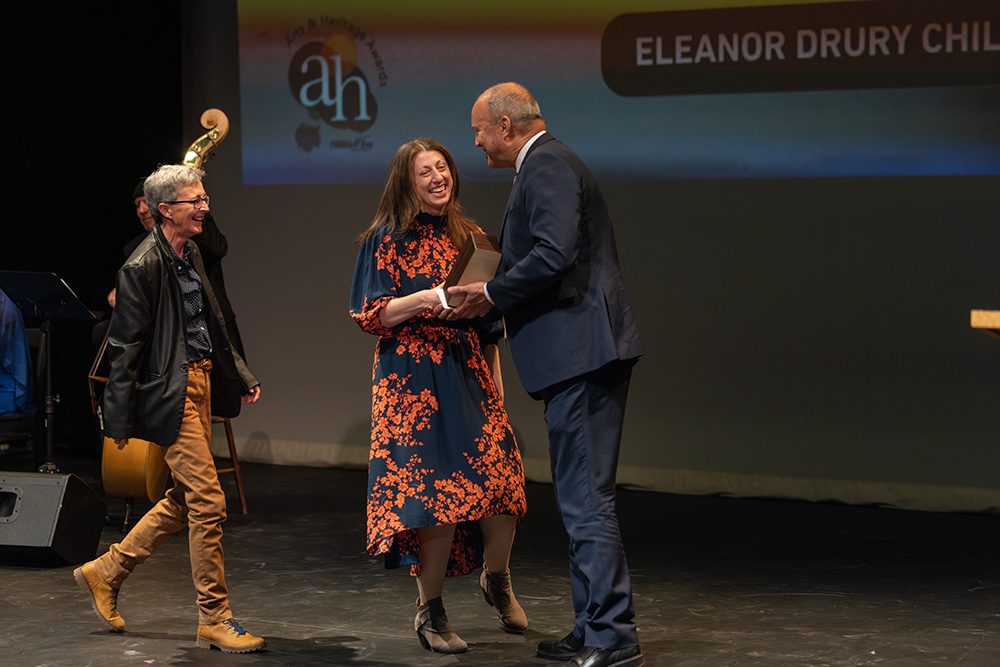 Woman in navy and orange floral dress receiving award handshake on stage from presenter