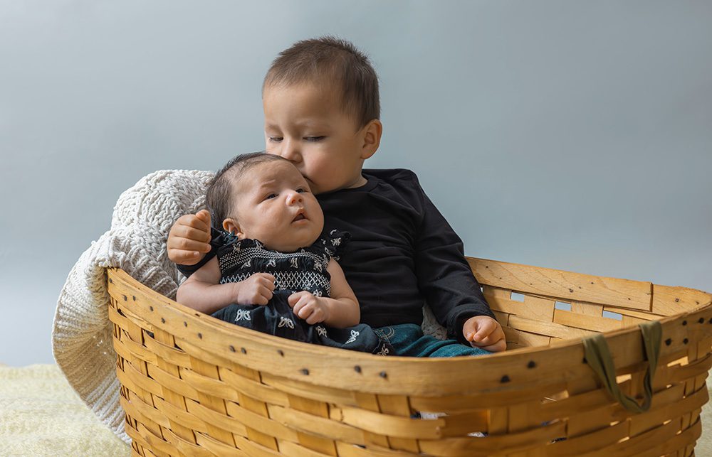 Toddler holding newborn baby in wicker basket studio sibling portrait