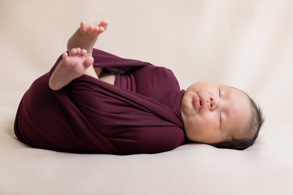 Newborn baby in burgundy swaddle with feet raised studio portrait