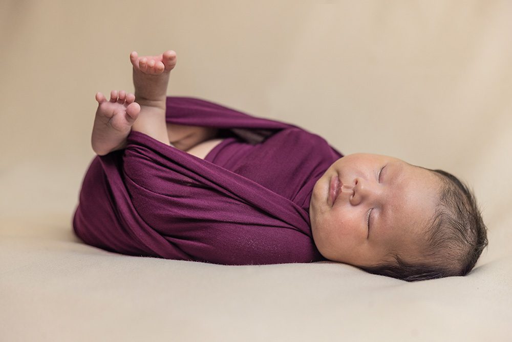 Swaddled newborn baby in burgundy wrap on cream background
