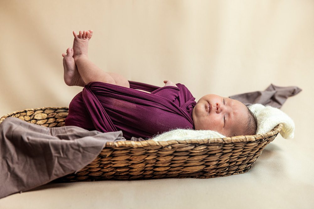 Newborn baby wrapped in burgundy swaddle in wicker basket feet up