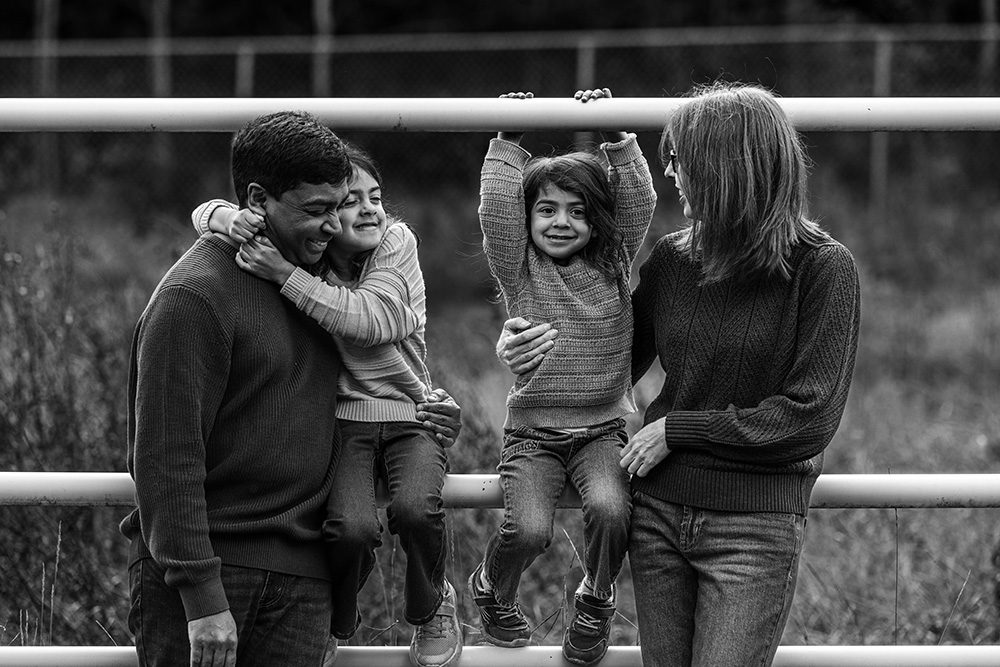 Black and white family portrait with playful children on white fence outdoor setting