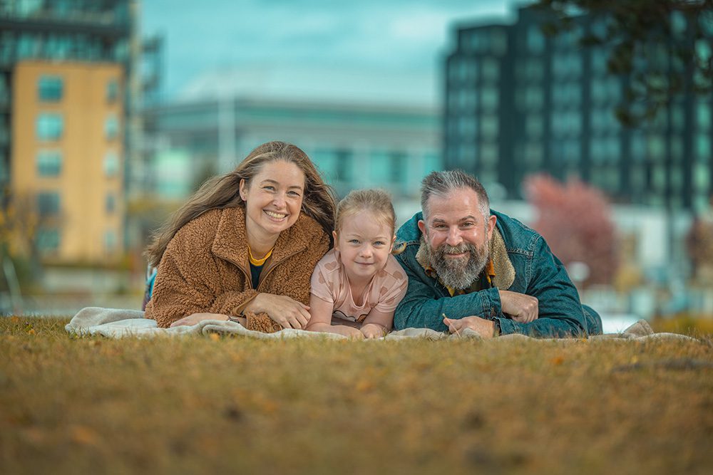 Family of three laying on stomachs on blanket smiling at camera with urban waterfront background