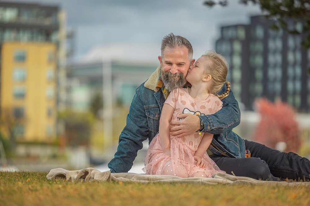 Father smiling while young daughter kisses his cheek during outdoor family photo session
