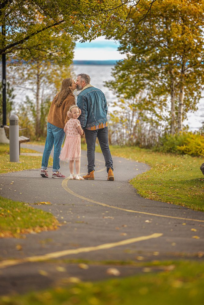 Parents kissing while young daughter stands between them on autumn pathway near water