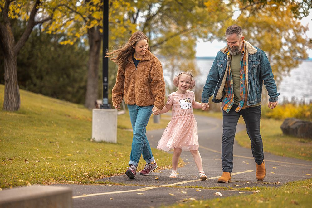 Family of three holding hands walking on autumn path with golden trees and water view