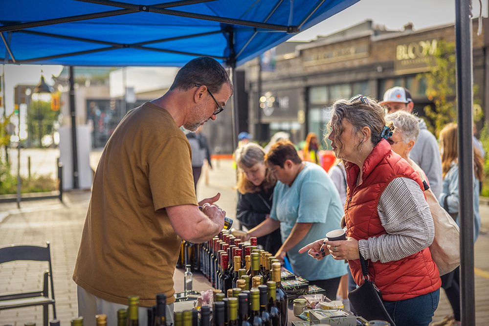 A vendor speaks with a customer while rows of bottled products are displayed on a table at an outdoor market.