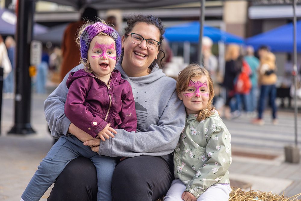 An adult sits smiling with two young children who have face paint at an outdoor market.