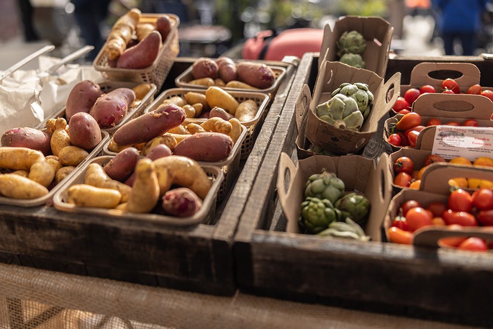 Assorted fresh vegetables, including potatoes, cherry tomatoes, and artichokes, displayed in wooden crates at an outdoor farmers’ market.