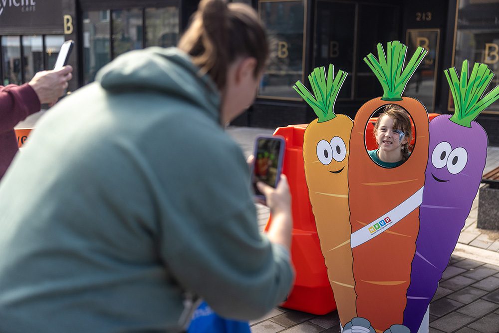 A child smiling through a carrot-shaped photo cutout while an adult takes a picture at an outdoor community event.