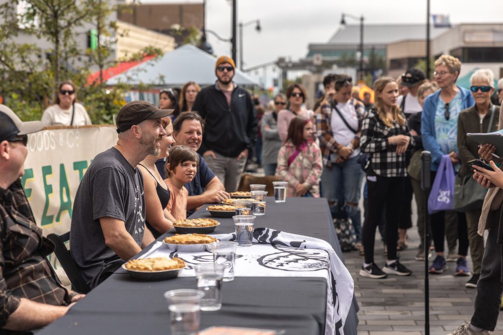 Several people sit behind a long table with pies while a crowd watches at an outdoor event.