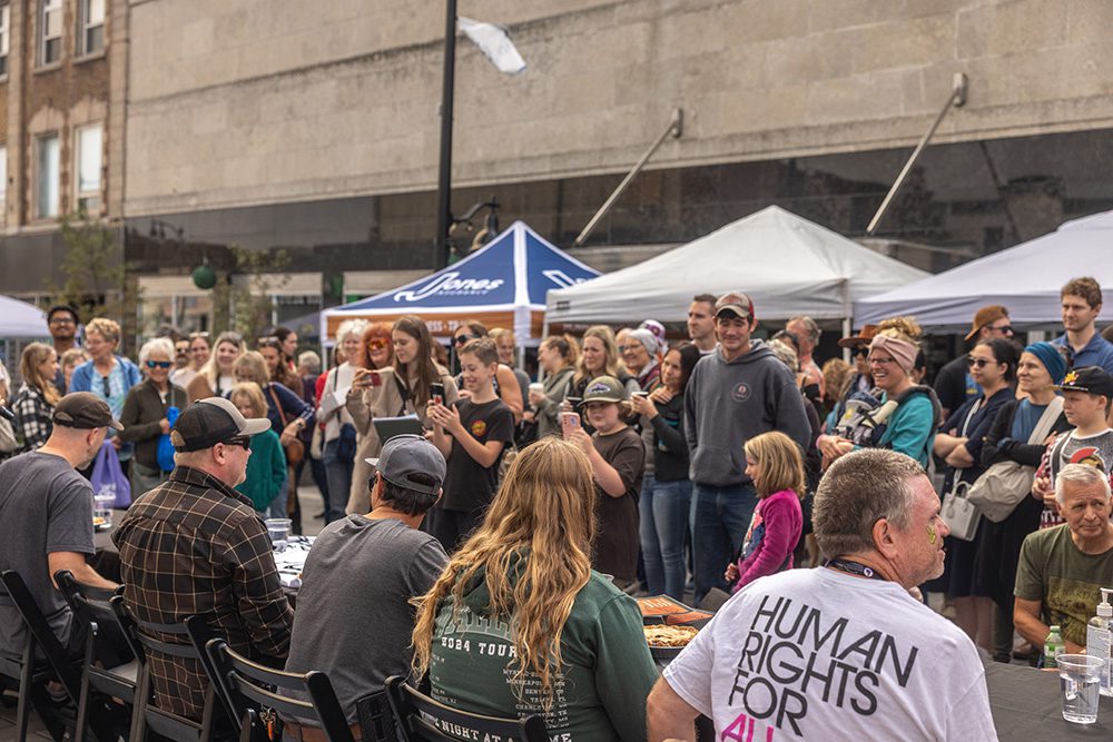 A large crowd stands around tables at an outdoor market event in a downtown setting.