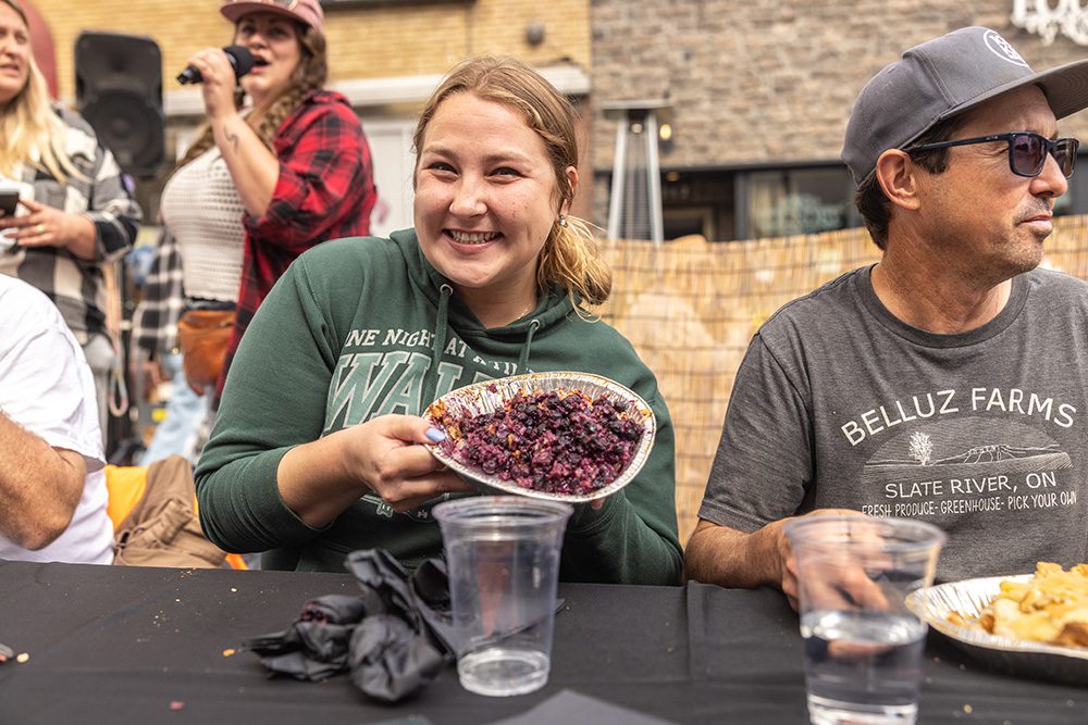 A smiling person holds a dish of food while seated at a table during an outdoor market event.