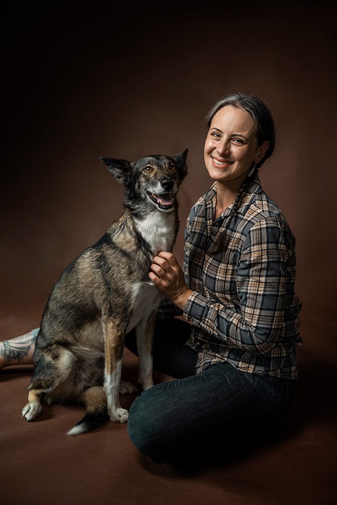Woman in plaid shirt sitting with medium-sized dog on brown studio backdrop