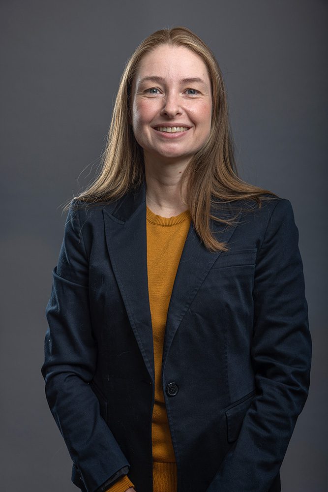 Professional woman in mustard sweater and navy blazer smiling against gray background for Professional Headshots Thunder Bay