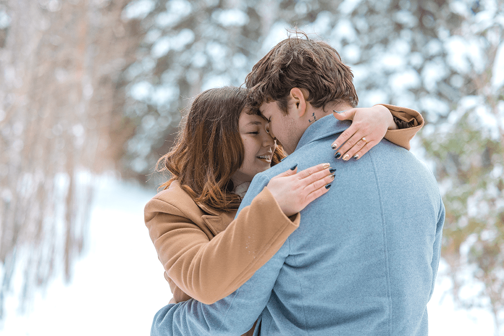 A couple embraces closely in a snowy forest, foreheads touching.