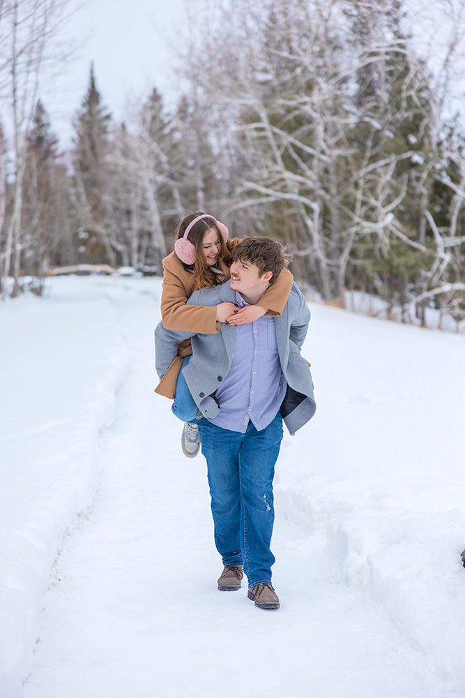 One partner gives the other a piggyback ride on a snowy path, both smiling.