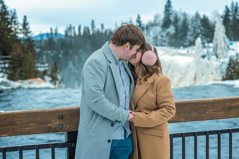 A couple stands close together by a wooden railing with Kakabeka Falls behind them in winter.