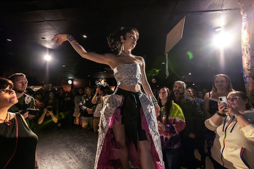 Model in newspaper-print outfit walking runway with audience watching