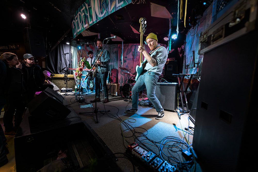 Live band performing on stage with guitarist in beanie under stage lights