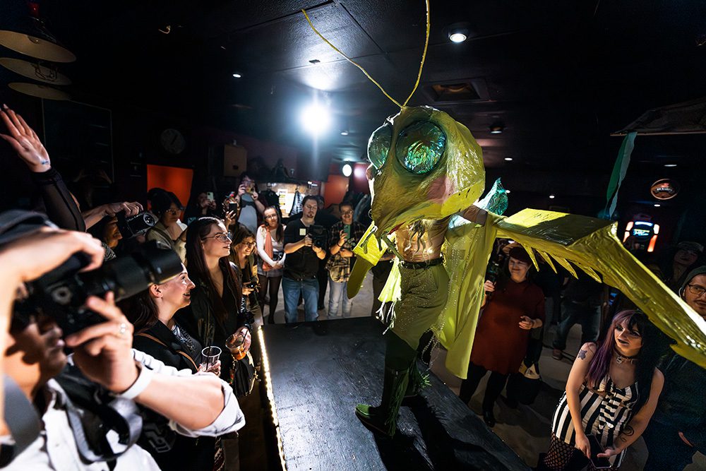 Dramatic yellow sculptural costume with insect motif surrounded by audience