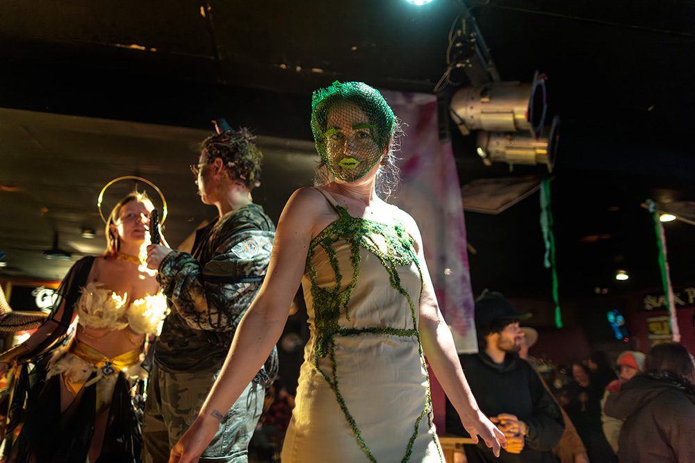 Model in green moss-themed costume with veil on runway at fashion show