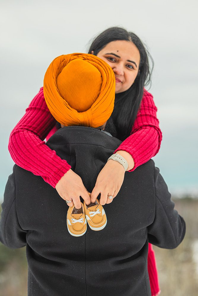 Pregnant woman being embraced by partner holding baby shoes during winter maternity session