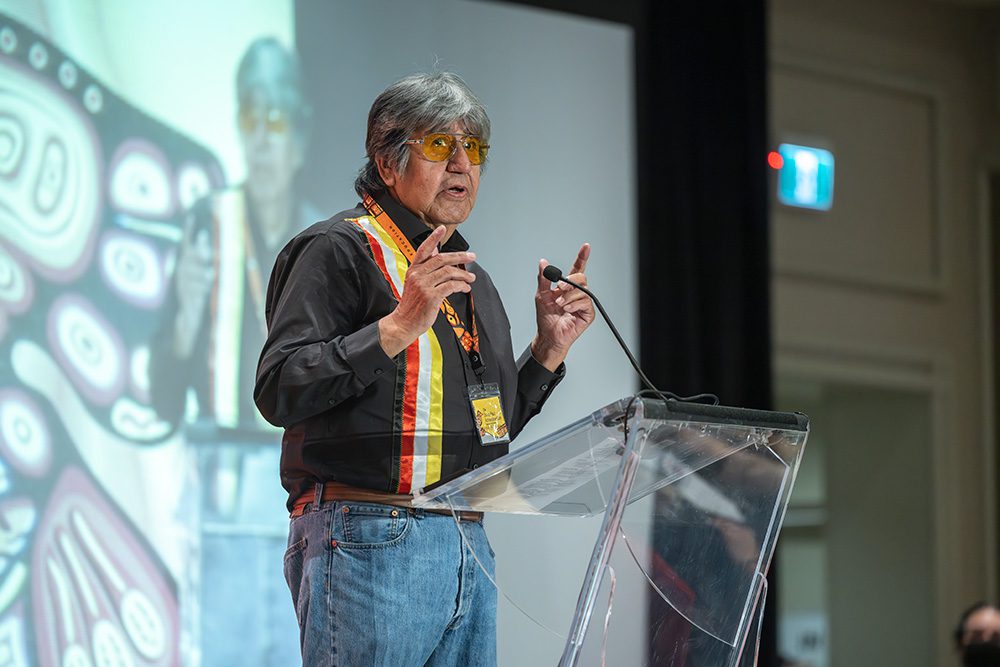 Indigenous elder speaking at clear podium with projected visuals behind