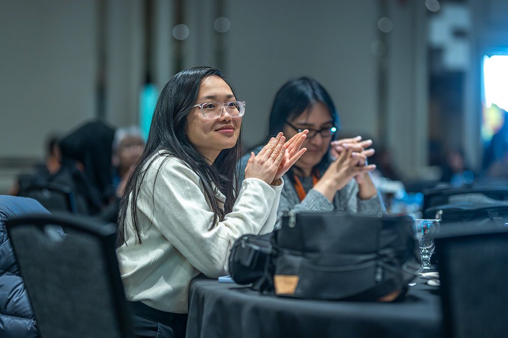Woman in white blazer applauding and smiling during conference presentation