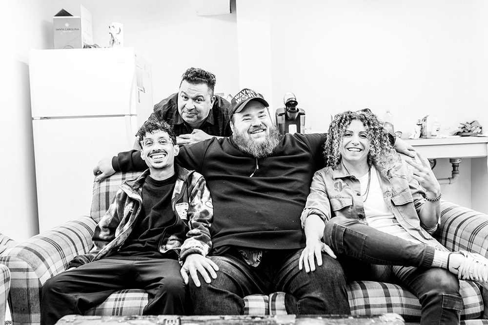 Black and white photo of four comedians sitting together on plaid couch backstage