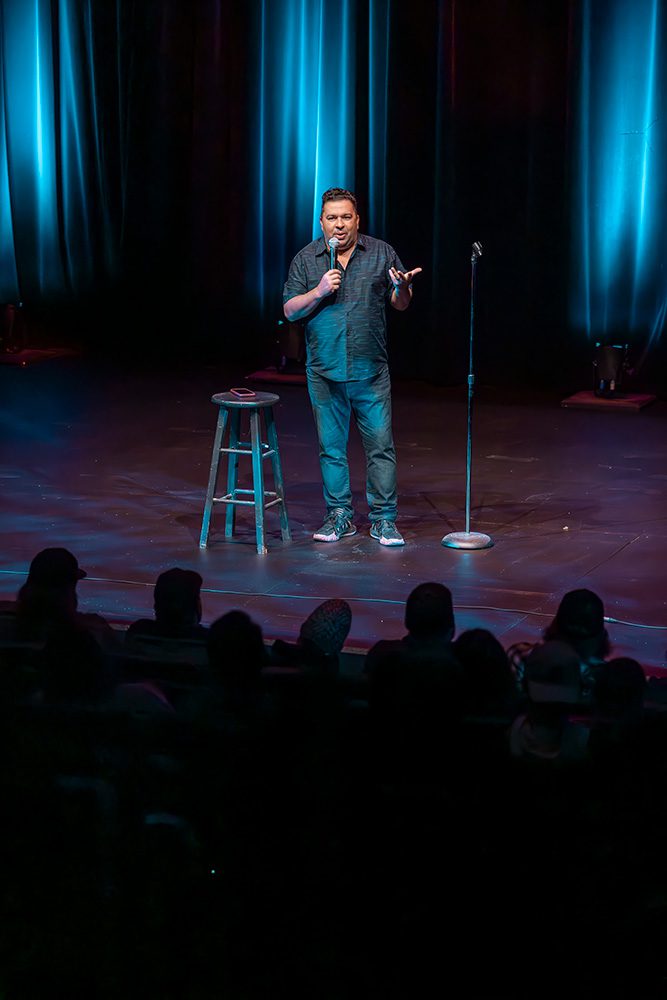 Comedian performing on stage under blue spotlight with audience silhouettes