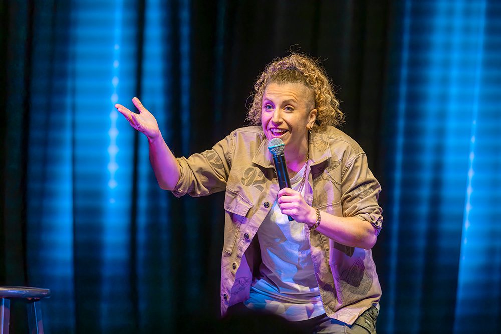 Female comedian in tan jacket gesturing with hand during stand-up performance