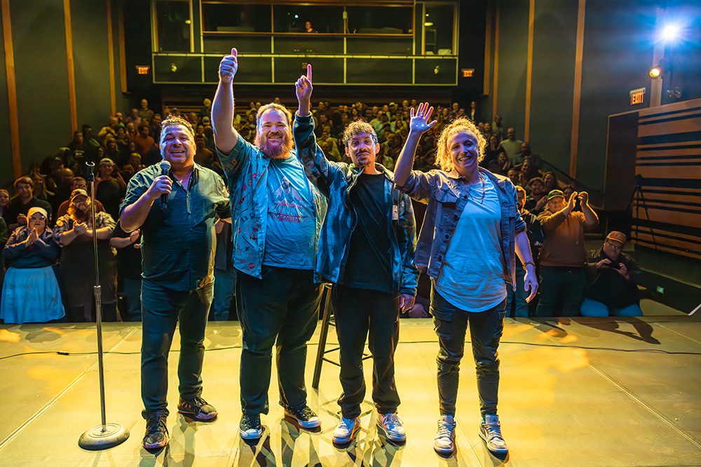 Four comedians on stage with arms raised celebrating packed comedy show audience
