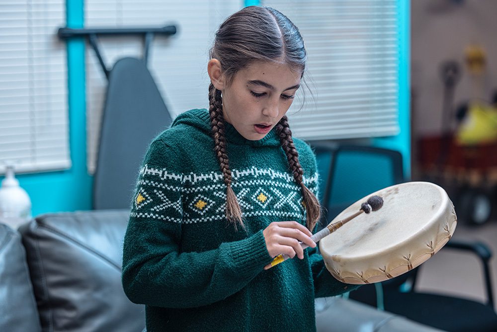 Young Indigenous girl with braids playing traditional hand drum at Anishnawbe Mushkiki cultural program