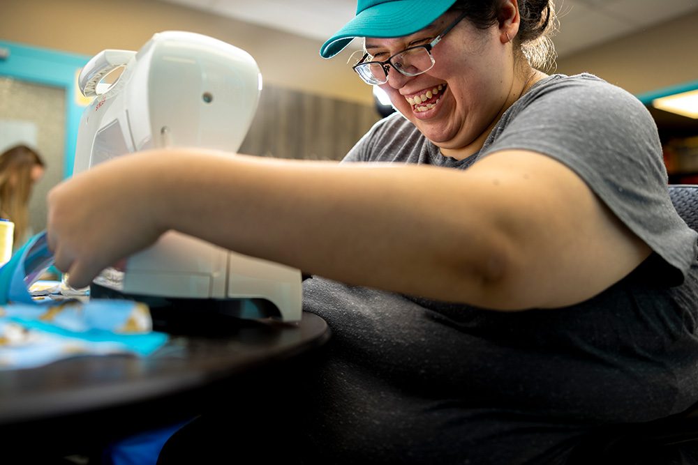 Woman in grey shirt and teal cap smiling while sewing at community workshop