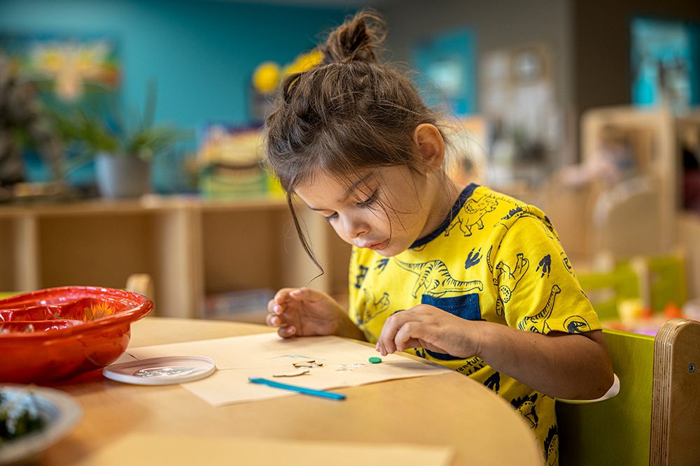 Young girl in yellow shirt concentrating on craft activity at community table