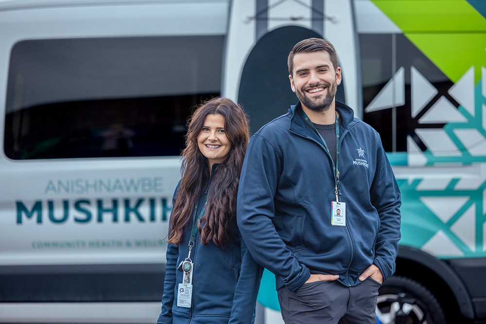Two Anishnawbe Mushkiki staff members smiling outdoors beside branded medical transport van