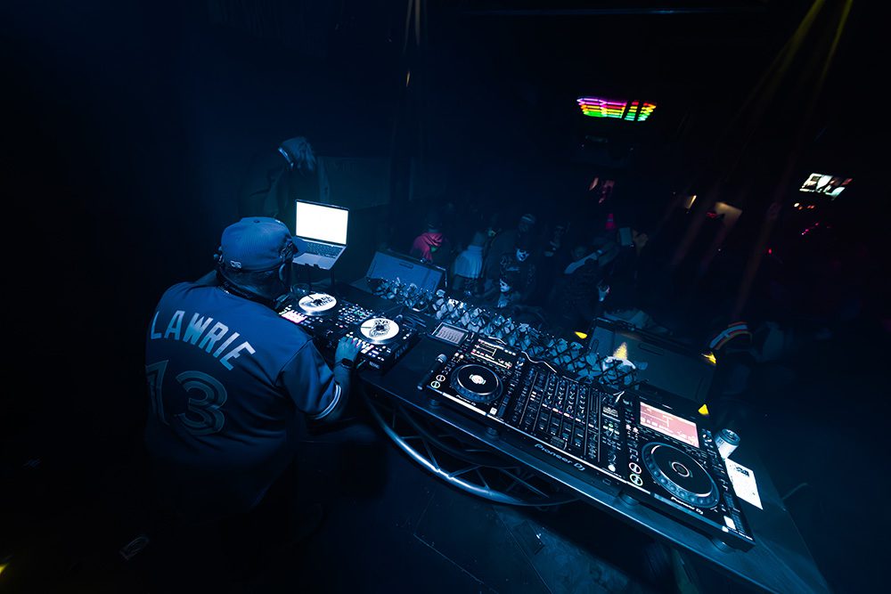 DJ performing with turntables and laptop under blue lighting at nightclub
