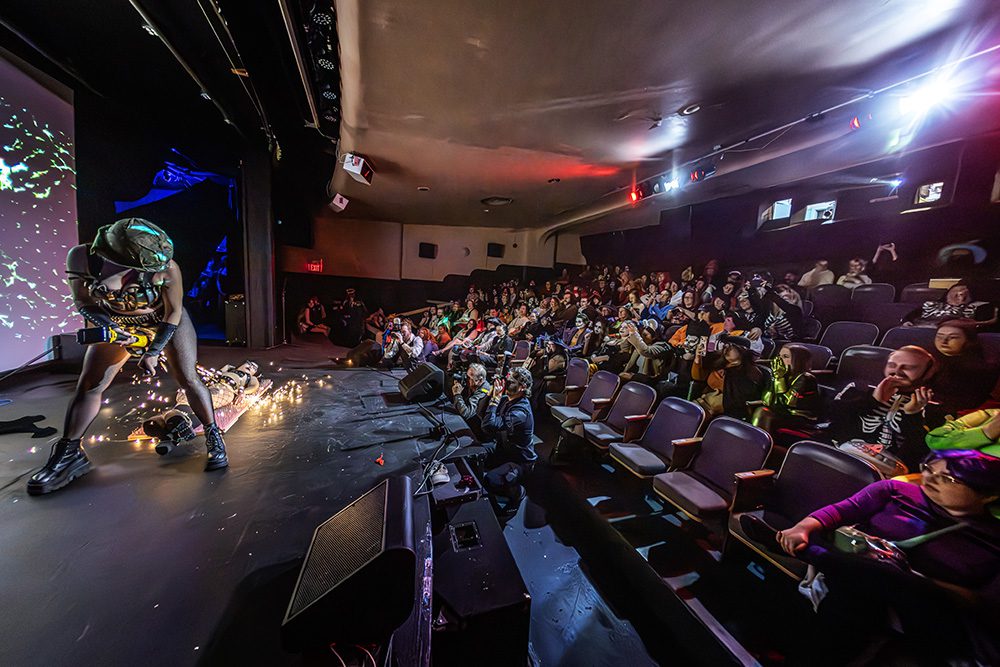 Performer on stage in theater-style venue with seated audience and atmospheric lighting during Halloween event
