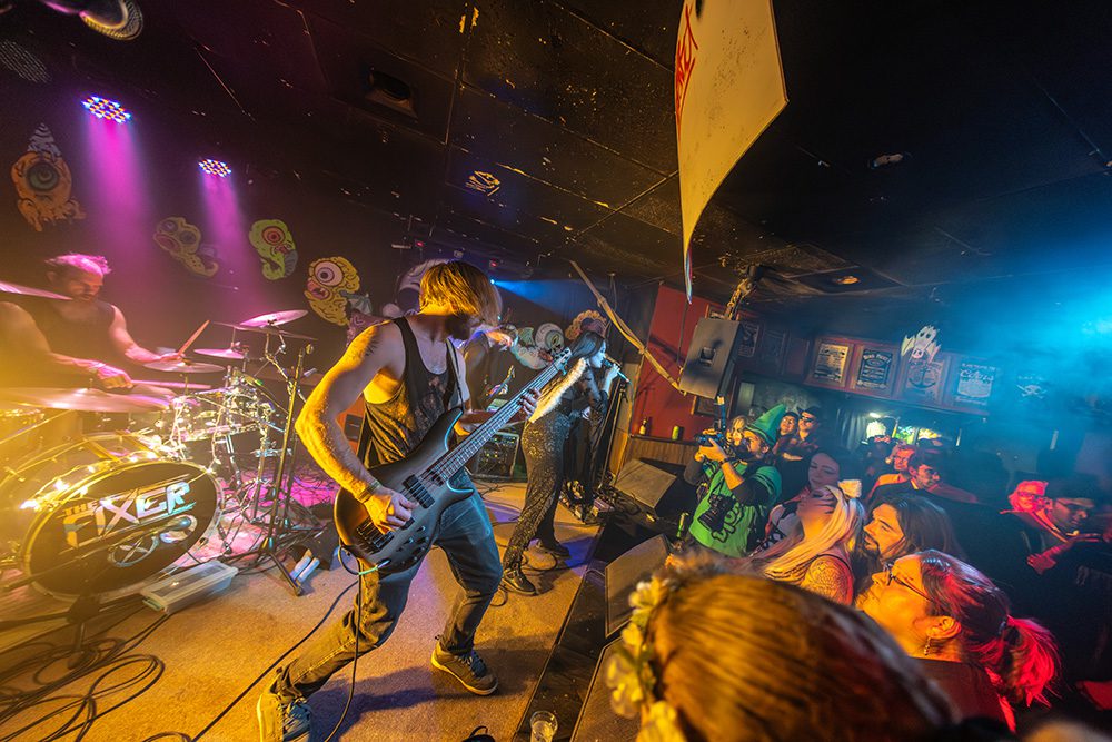 Rock band performing on intimate stage with bass guitarist in foreground and engaged audience watching at close range
