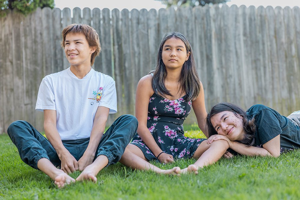 Three teenage siblings sitting and lying casually on grass in backyard laughing together