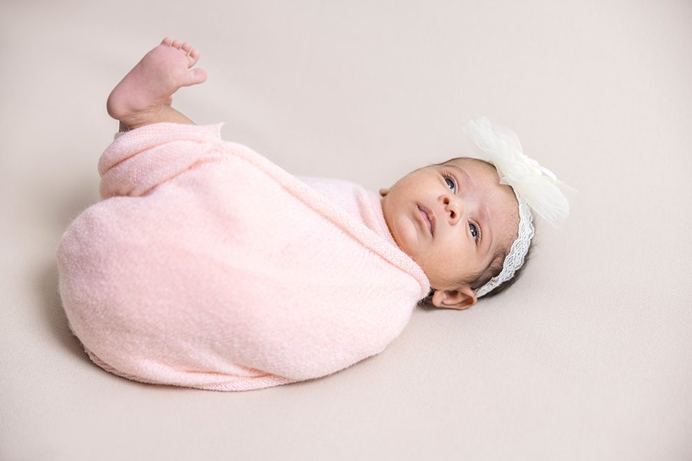 Newborn baby in soft pink wrap with white bow lying on side on neutral background