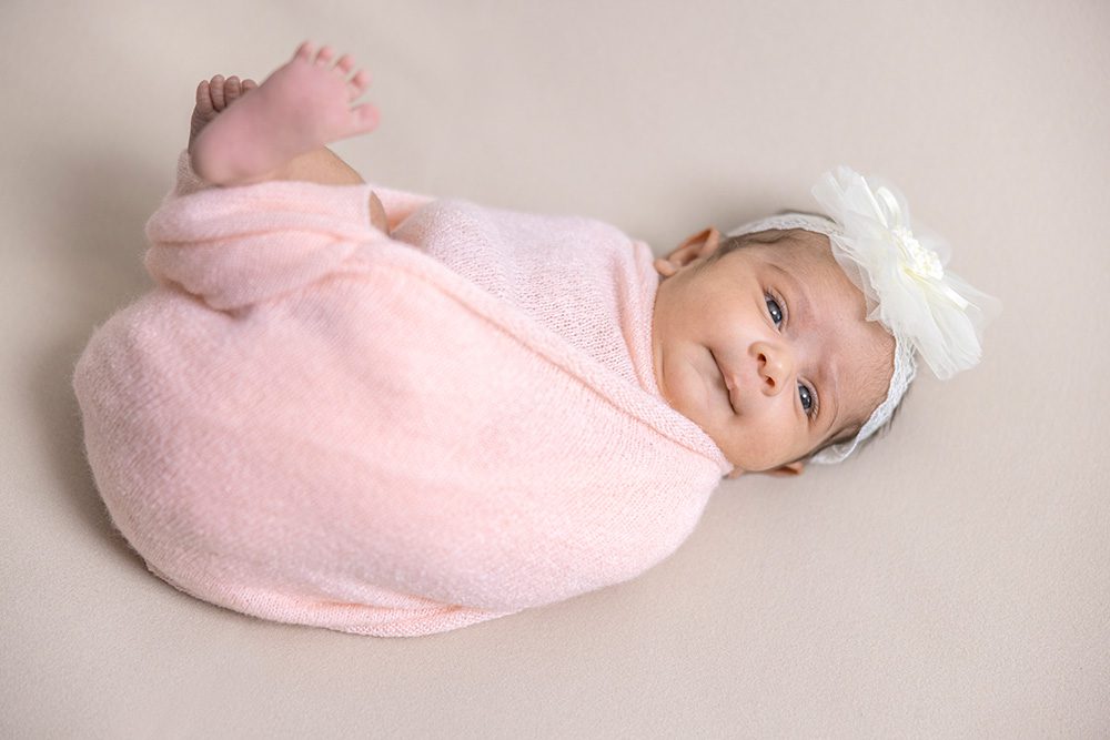 Newborn baby in pink wrap with white flower bow headband lying on neutral backdrop