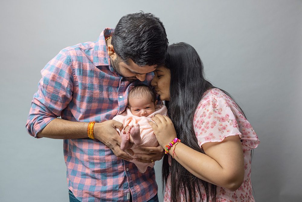 New parents in plaid shirts holding newborn baby together against gray background